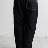 A person shown from the back wearing loose black denim jeans with contrast white stitching, a brown sweatshirt, and dark sneakers against a plain white wall.