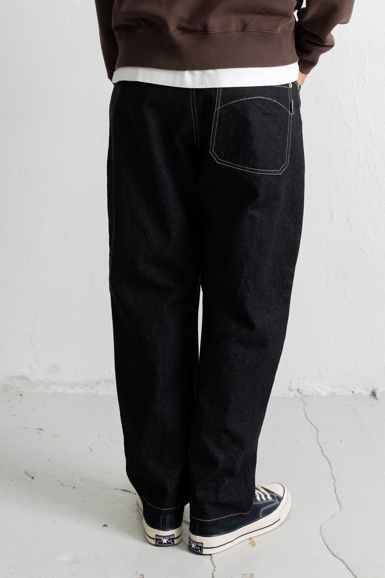 A person shown from the back wearing loose black denim jeans with contrast white stitching, a brown sweatshirt, and dark sneakers against a plain white wall.