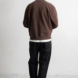 A man viewed from behind wearing a brown crewneck sweatshirt, black jeans, and black canvas sneakers, standing against a plain white wall.