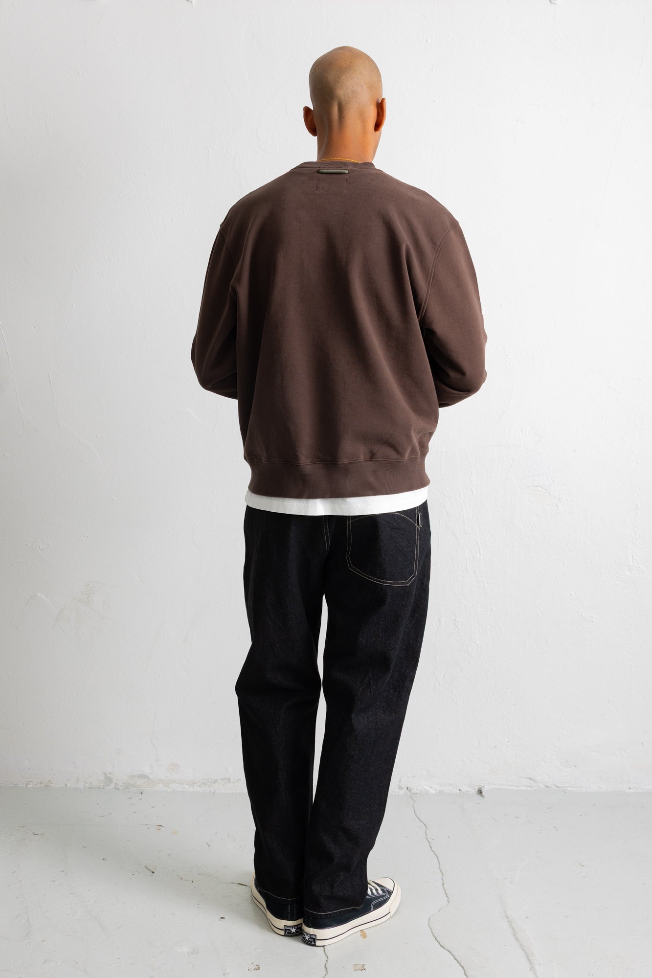 A man viewed from behind wearing a brown crewneck sweatshirt, black jeans, and black canvas sneakers, standing against a plain white wall.