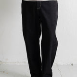 A person wearing loose black denim jeans, a brown sweatshirt, and black canvas sneakers, standing against a white wall.