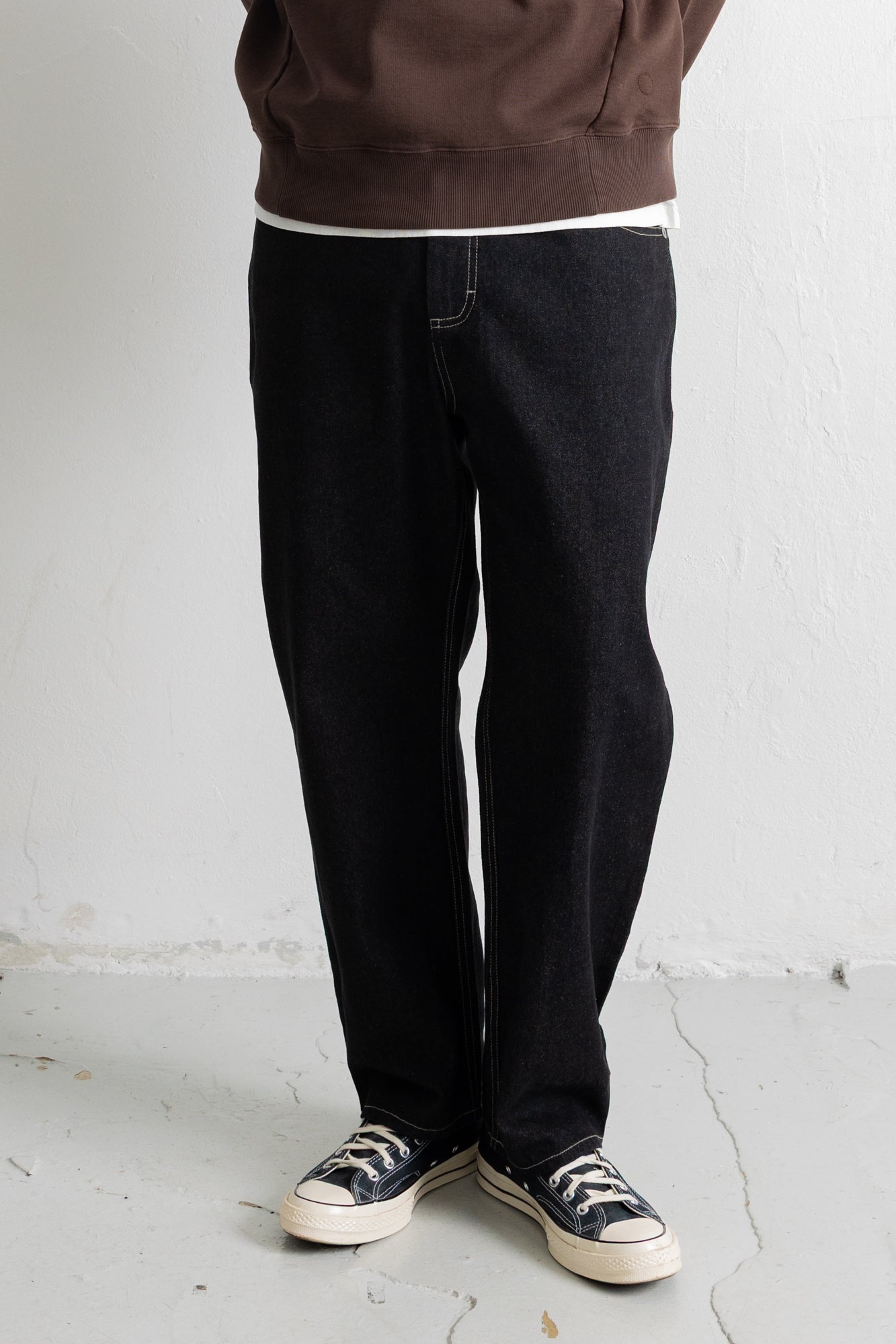 A person wearing loose black denim jeans, a brown sweatshirt, and black canvas sneakers, standing against a white wall.