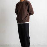 A man wearing a dark brown crewneck sweatshirt, black jeans, and black canvas sneakers, standing against a plain white wall.