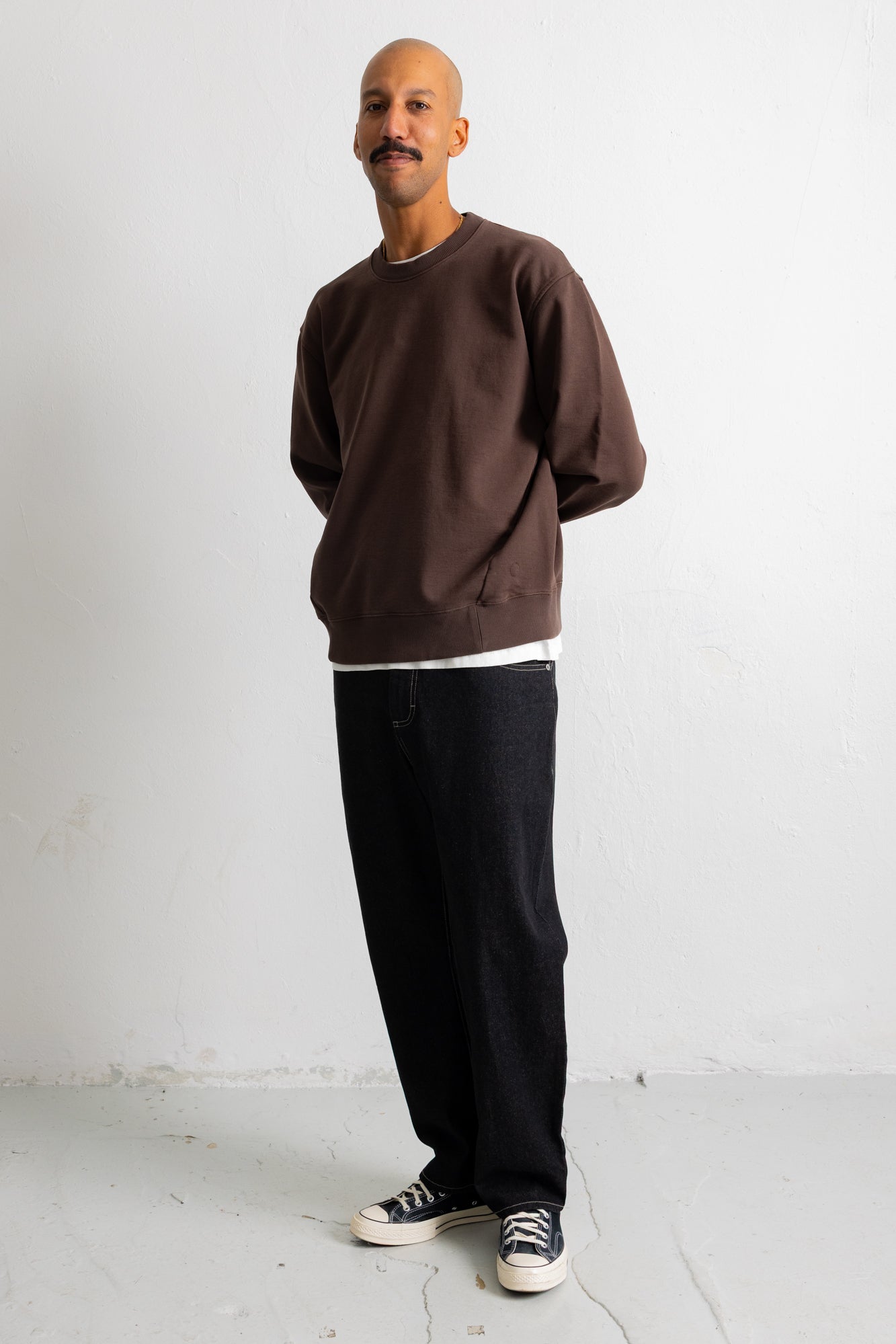 A man wearing a dark brown crewneck sweatshirt, black jeans, and black canvas sneakers, standing against a plain white wall.