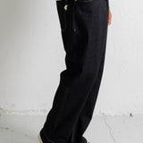 A person wearing loose black denim jeans with a brown sweatshirt and black canvas sneakers, standing against a light gray wall.
