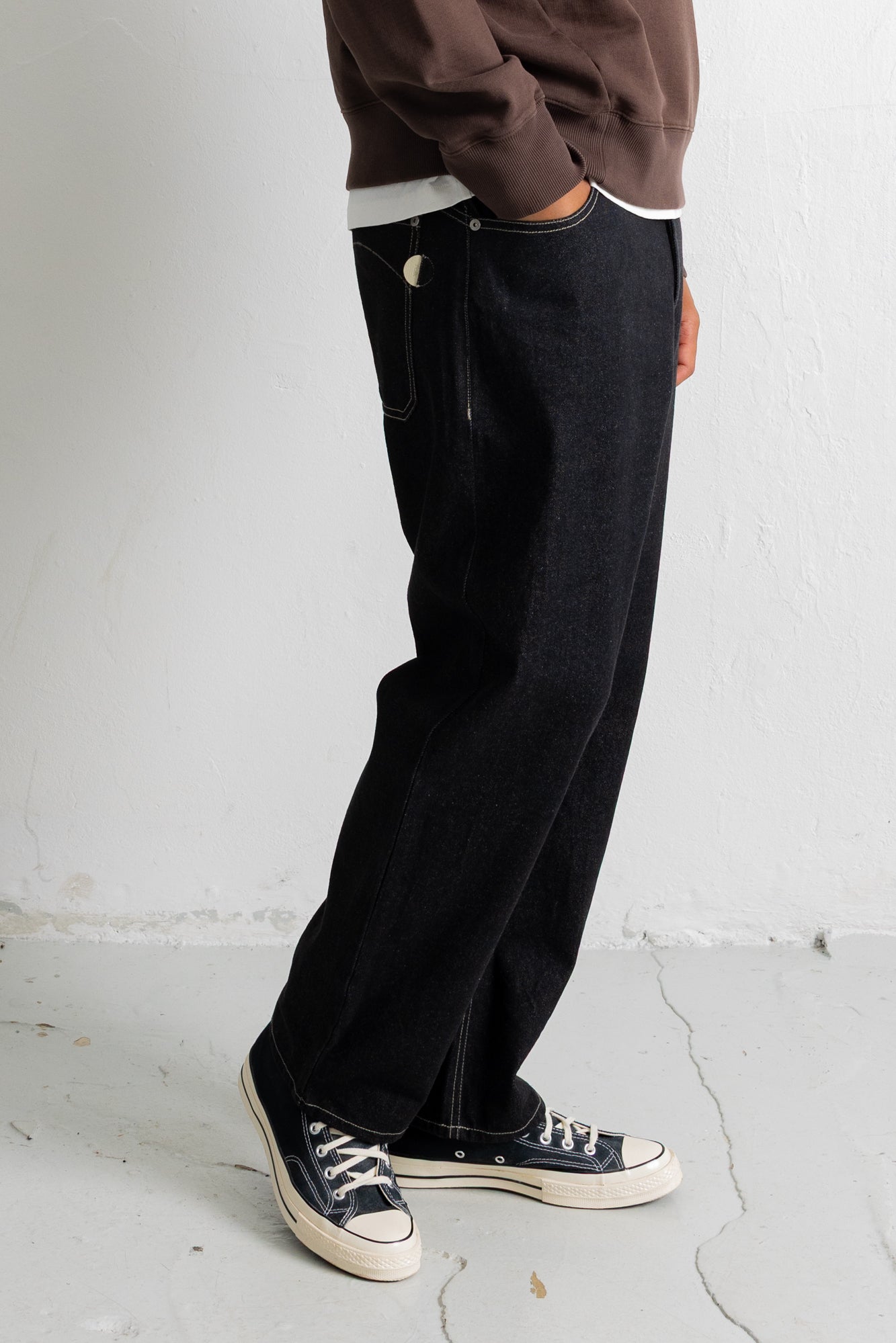 A person wearing loose black denim jeans with a brown sweatshirt and black canvas sneakers, standing against a light gray wall.