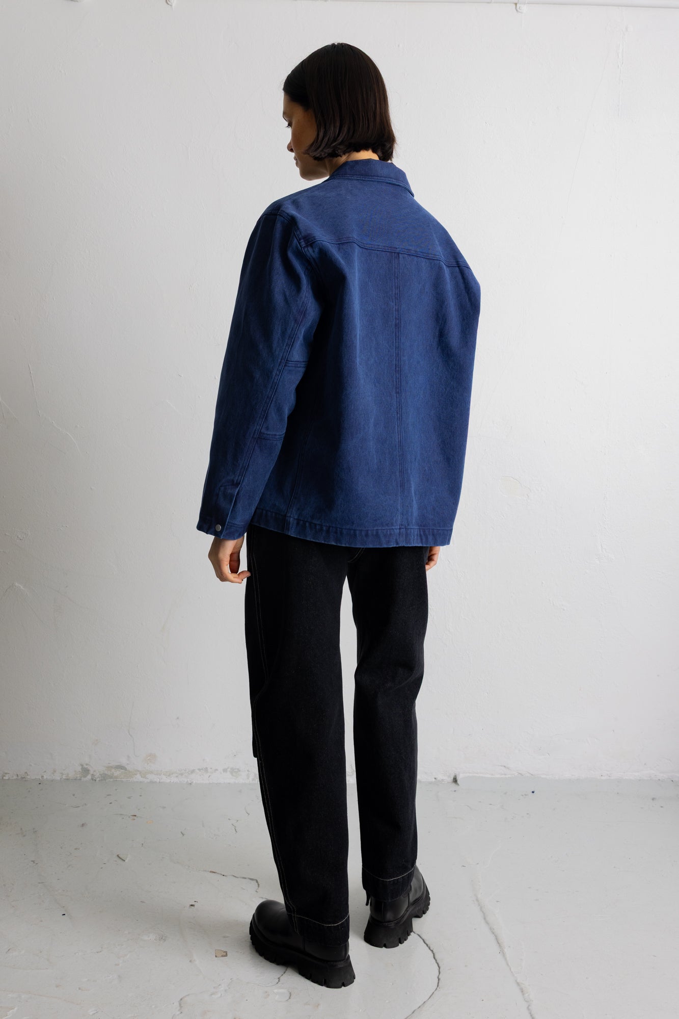 A person shown from the back wearing a blue denim-style jacket, black pants, and black boots in a plain white studio setting.
