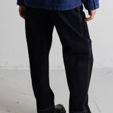 A person shown from the back wearing loose black jeans with contrast stitching, a dark blue jacket, and black leather boots on a light gray concrete floor.