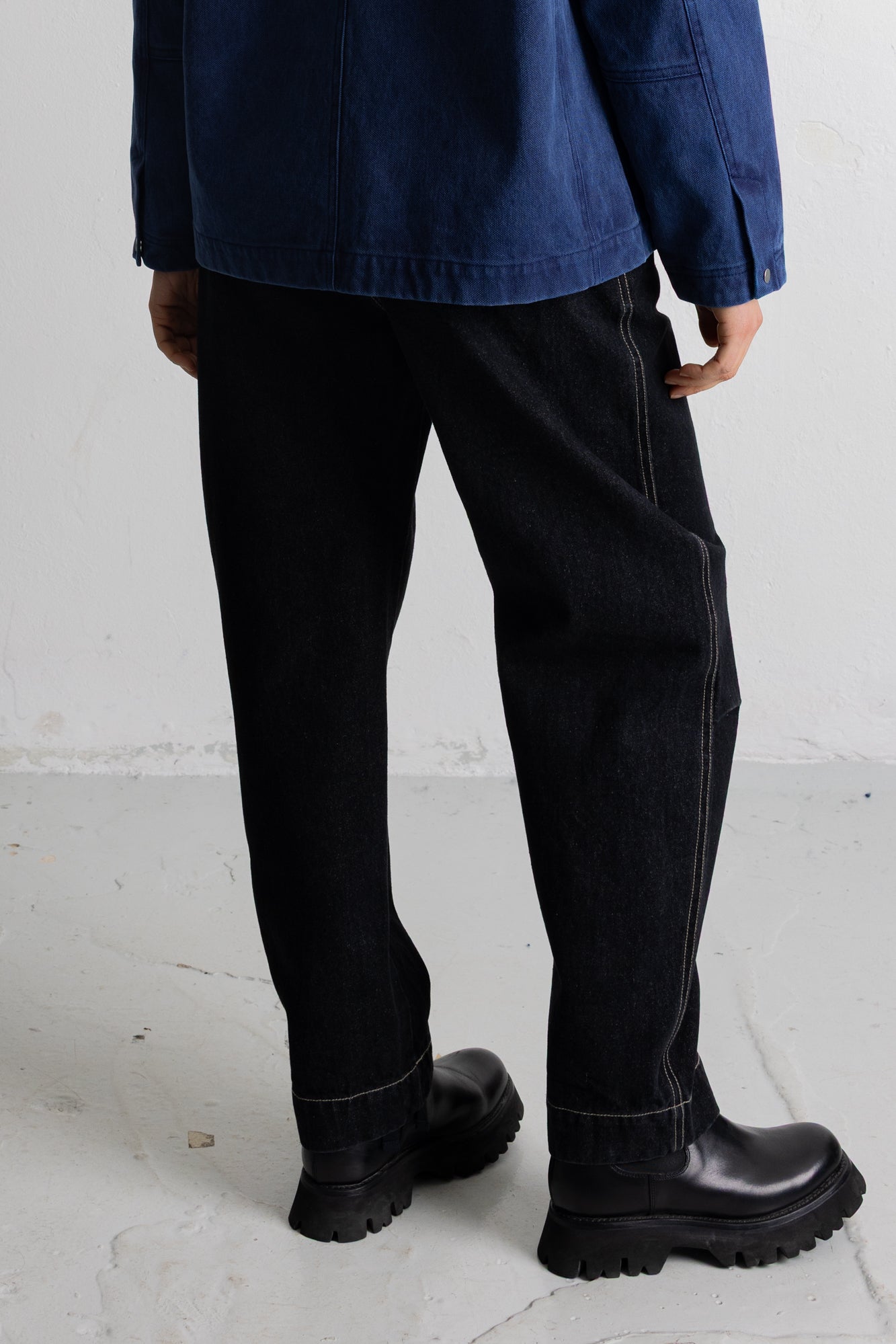 A person shown from the back wearing loose black jeans with contrast stitching, a dark blue jacket, and black leather boots on a light gray concrete floor.
