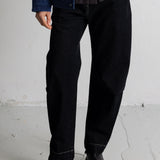 A person wearing black straight-leg denim jeans, a dark striped shirt, a blue denim jacket, and black leather boots, standing against a light gray concrete wall.