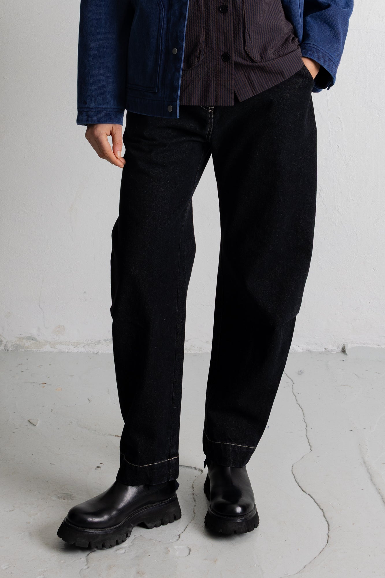 A person wearing black straight-leg denim jeans, a dark striped shirt, a blue denim jacket, and black leather boots, standing against a light gray concrete wall.