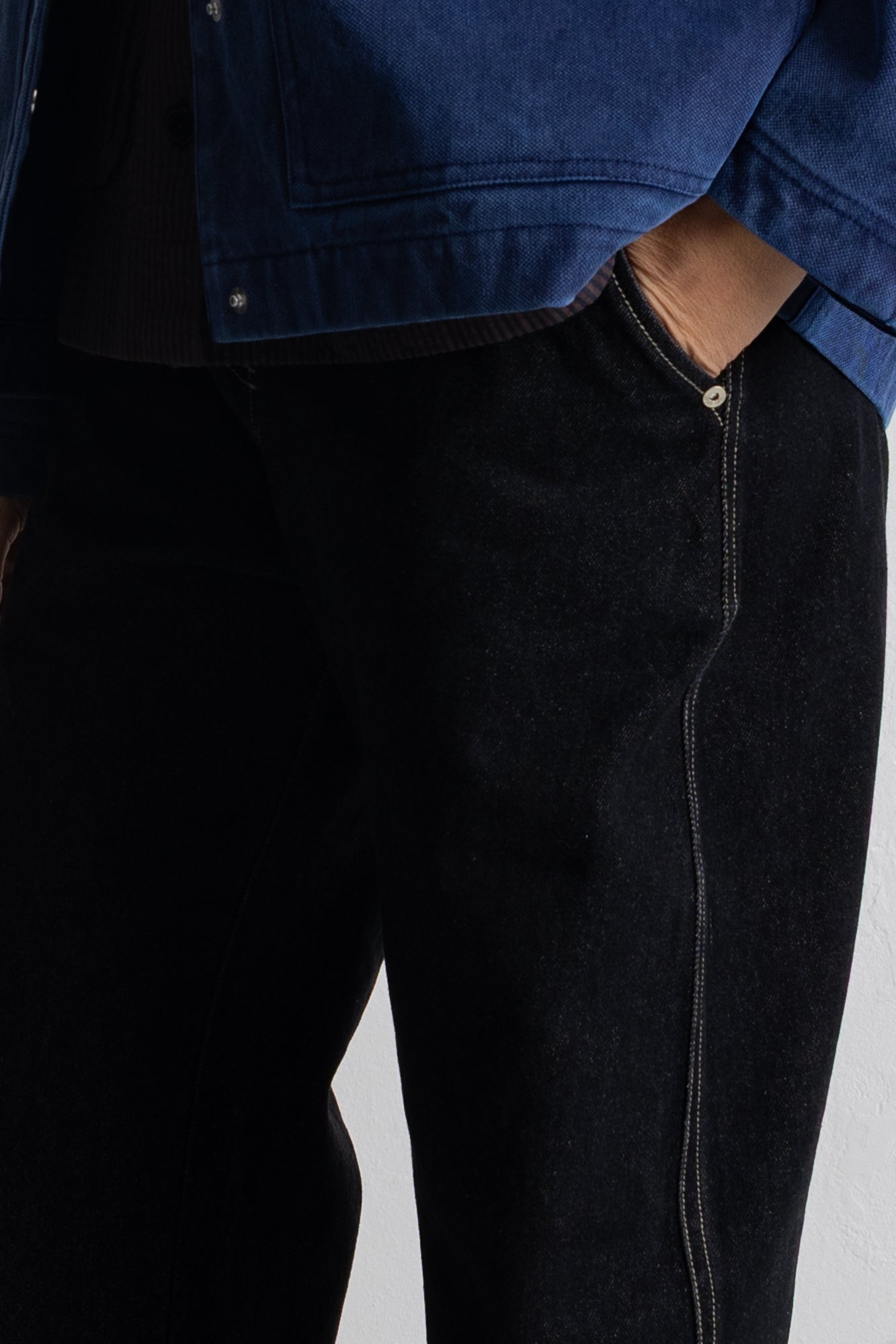 Close-up of a person wearing dark indigo denim jeans with contrast stitching and a blue denim jacket partially visible at the top.