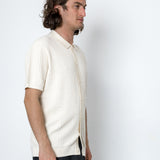 Man wearing a white ribbed knit shirt and dark pants against a white background