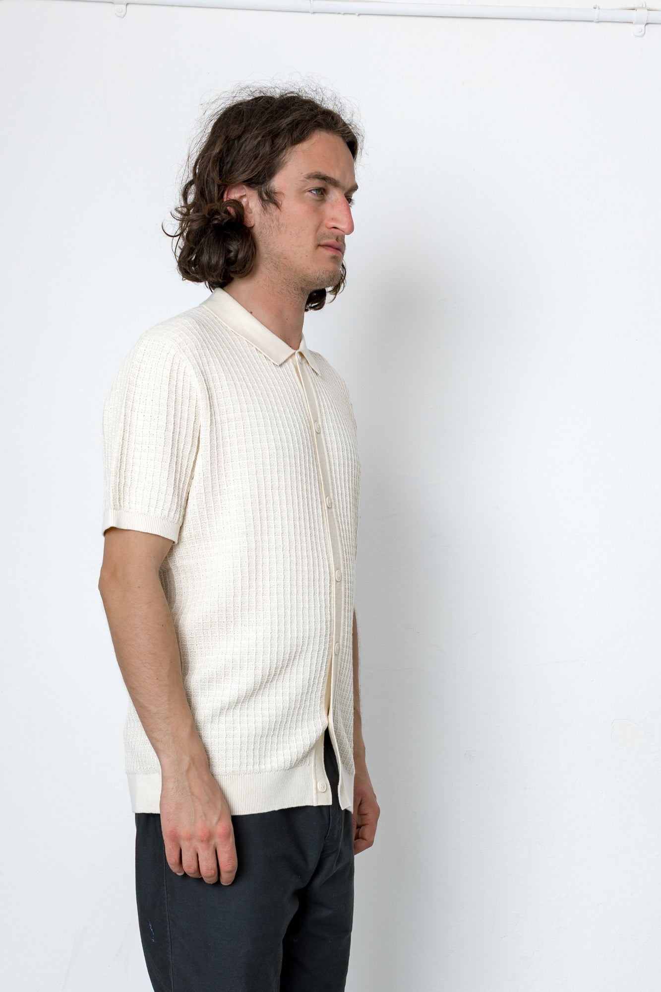 Man wearing a white ribbed knit shirt and dark pants against a white background