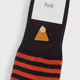 A single black crew sock with orange stripes near the cuff, featuring a small tan leather triangle charm with a button, shown with a white paper tag printed with the brand name 'Folk'.
