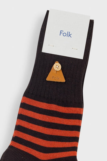 A single black crew sock with orange stripes near the cuff, featuring a small tan leather triangle charm with a button, shown with a white paper tag printed with the brand name 'Folk'.