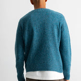 A man viewed from behind wearing a teal blue knit sweater over a white t-shirt and brown pants, against a plain white studio background.