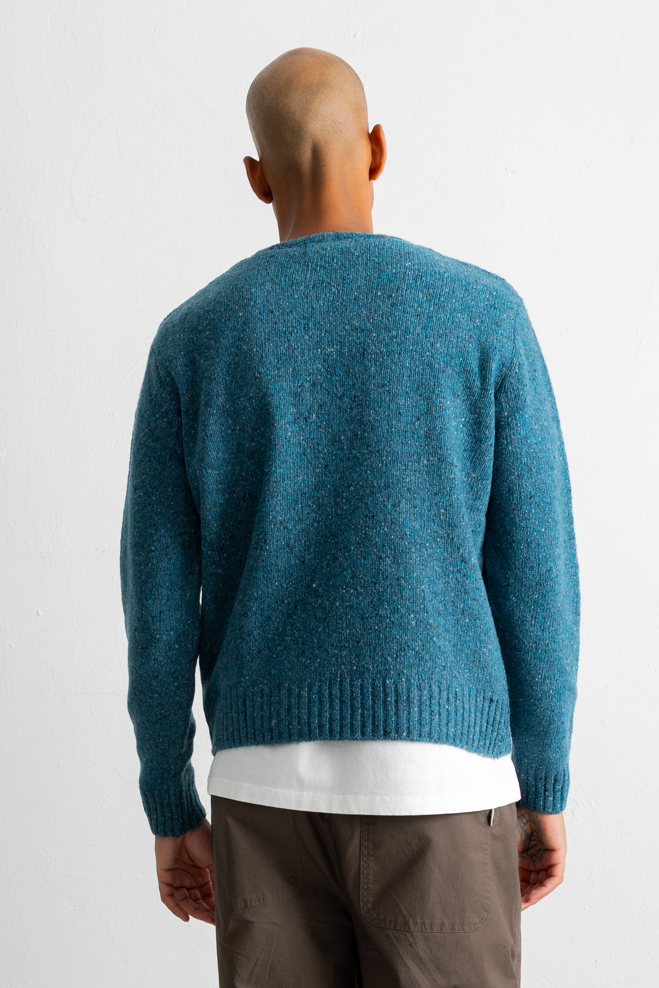 A man viewed from behind wearing a teal blue knit sweater over a white t-shirt and brown pants, against a plain white studio background.