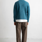A man viewed from behind wearing a teal blue knit sweater over a white t-shirt and brown trousers, standing against a plain white wall.