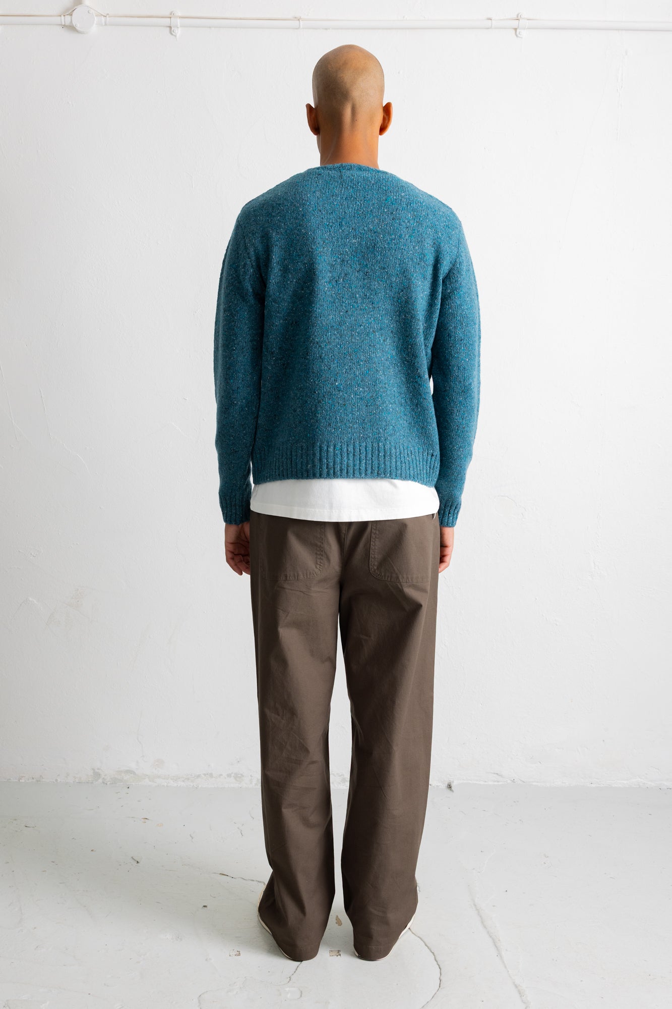 A man viewed from behind wearing a teal blue knit sweater over a white t-shirt and brown trousers, standing against a plain white wall.