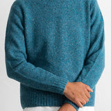 A man wearing a teal blue heathered knit crewneck sweater, styled with a white undershirt and brown trousers, against a plain white wall.