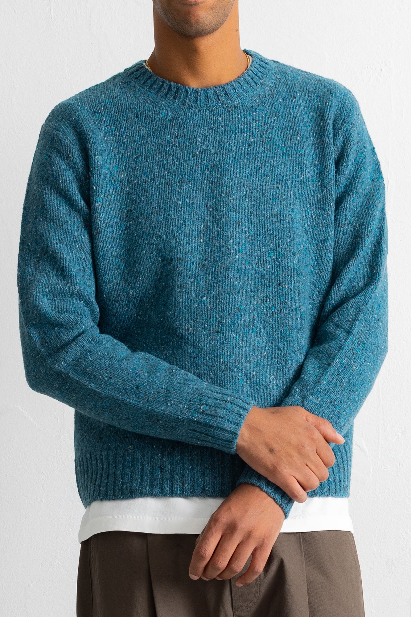 A man wearing a teal blue heathered knit crewneck sweater, styled with a white undershirt and brown trousers, against a plain white wall.