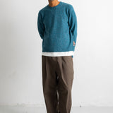 A man wearing a teal blue knit crewneck sweater, brown trousers, and black-and-white sneakers, standing against a plain white studio wall.