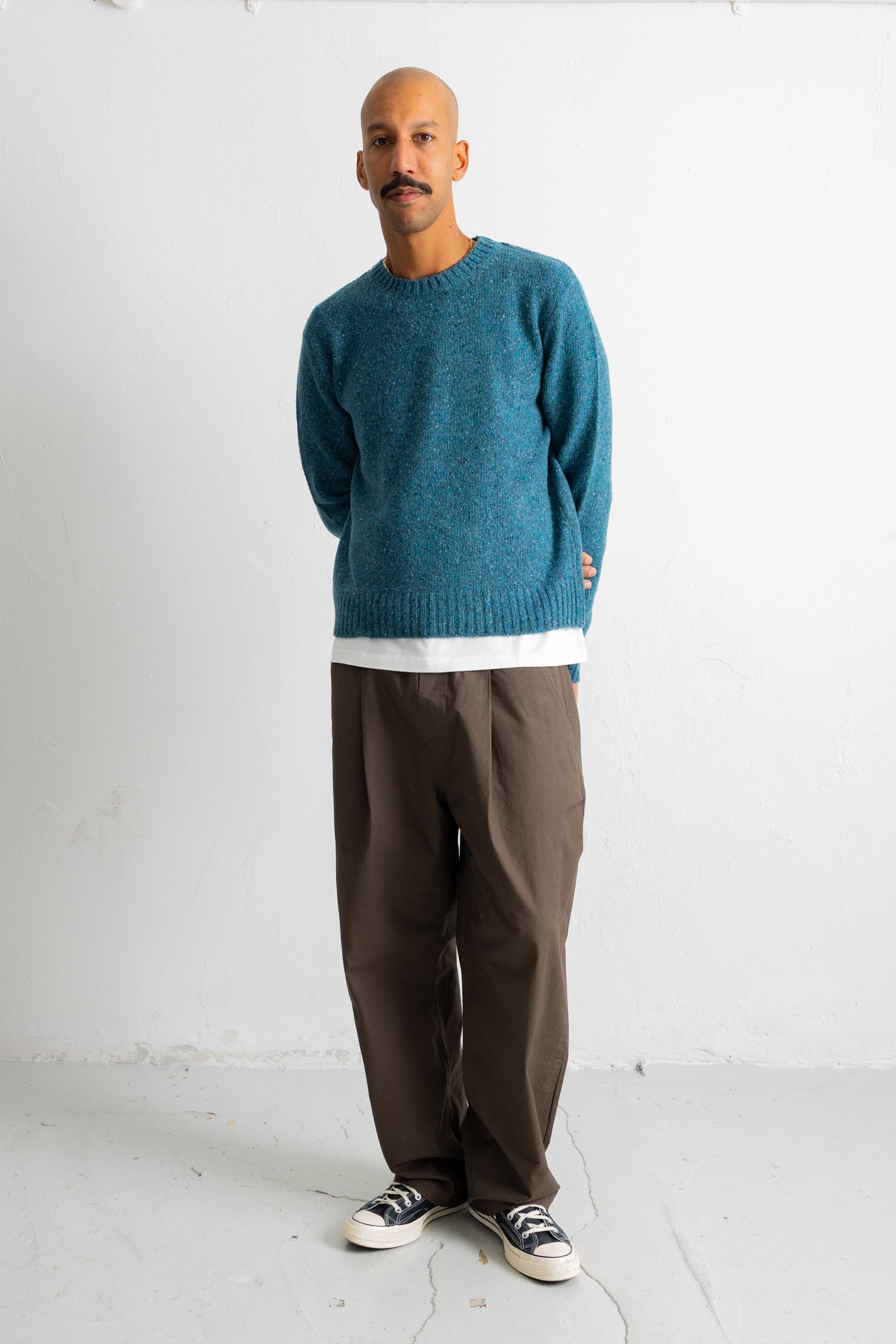 A man wearing a teal blue knit crewneck sweater, brown trousers, and black-and-white sneakers, standing against a plain white studio wall.