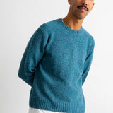 A man wearing a textured teal blue crewneck sweater over a white undershirt and brown pants, standing against a plain white wall.