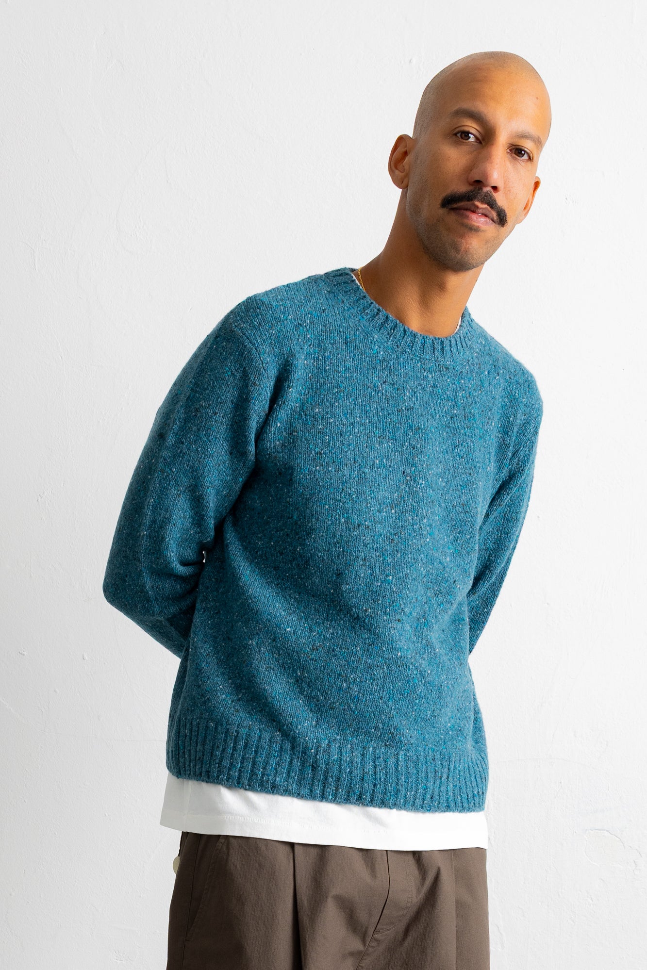 A man wearing a textured teal blue crewneck sweater over a white undershirt and brown pants, standing against a plain white wall.