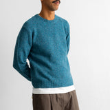 A man wearing a teal blue knit crewneck sweater over a white undershirt and brown trousers, standing against a plain white wall.