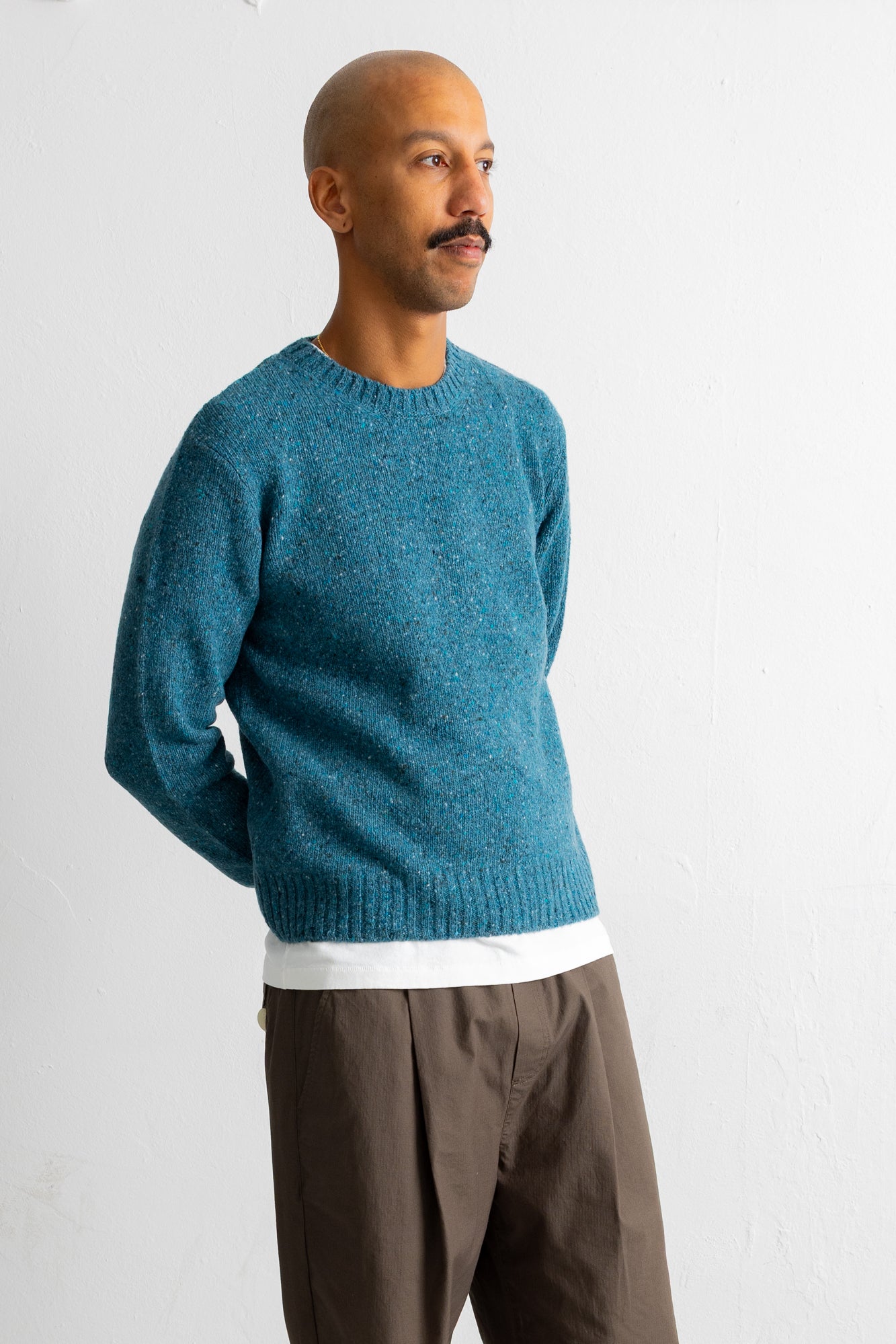 A man wearing a teal blue knit crewneck sweater over a white undershirt and brown trousers, standing against a plain white wall.