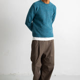 A man wearing a teal blue knit crewneck sweater, brown wide-leg trousers, and black canvas sneakers, standing against a white studio wall.