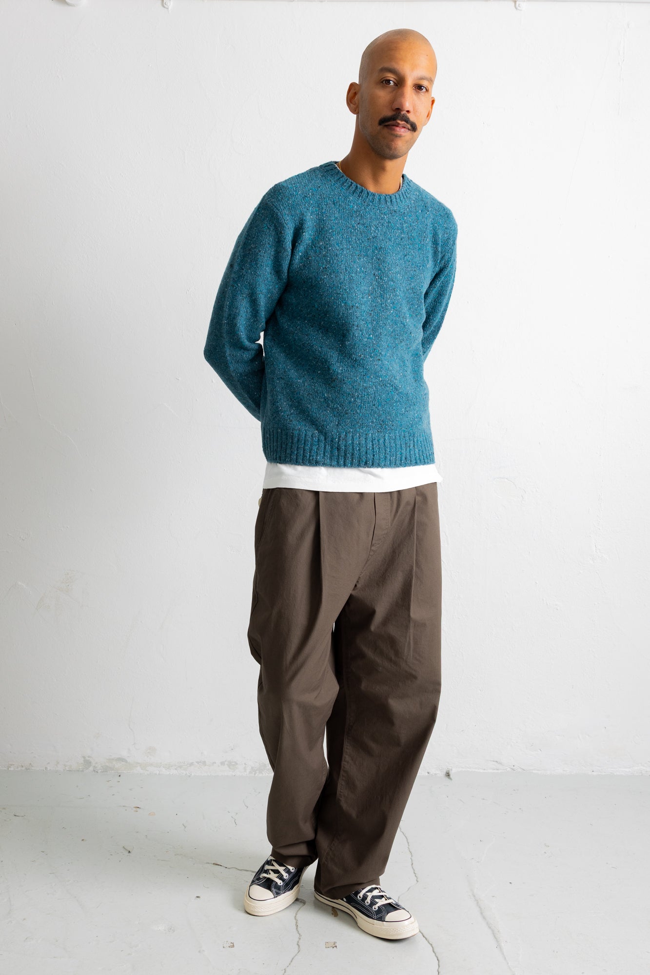 A man wearing a teal blue knit crewneck sweater, brown wide-leg trousers, and black canvas sneakers, standing against a white studio wall.