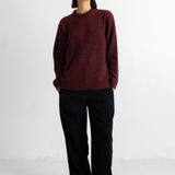 A woman stands against a plain white studio wall wearing a chunky knit crewneck sweater in deep burgundy, black wide-leg trousers, and black pointed flats.