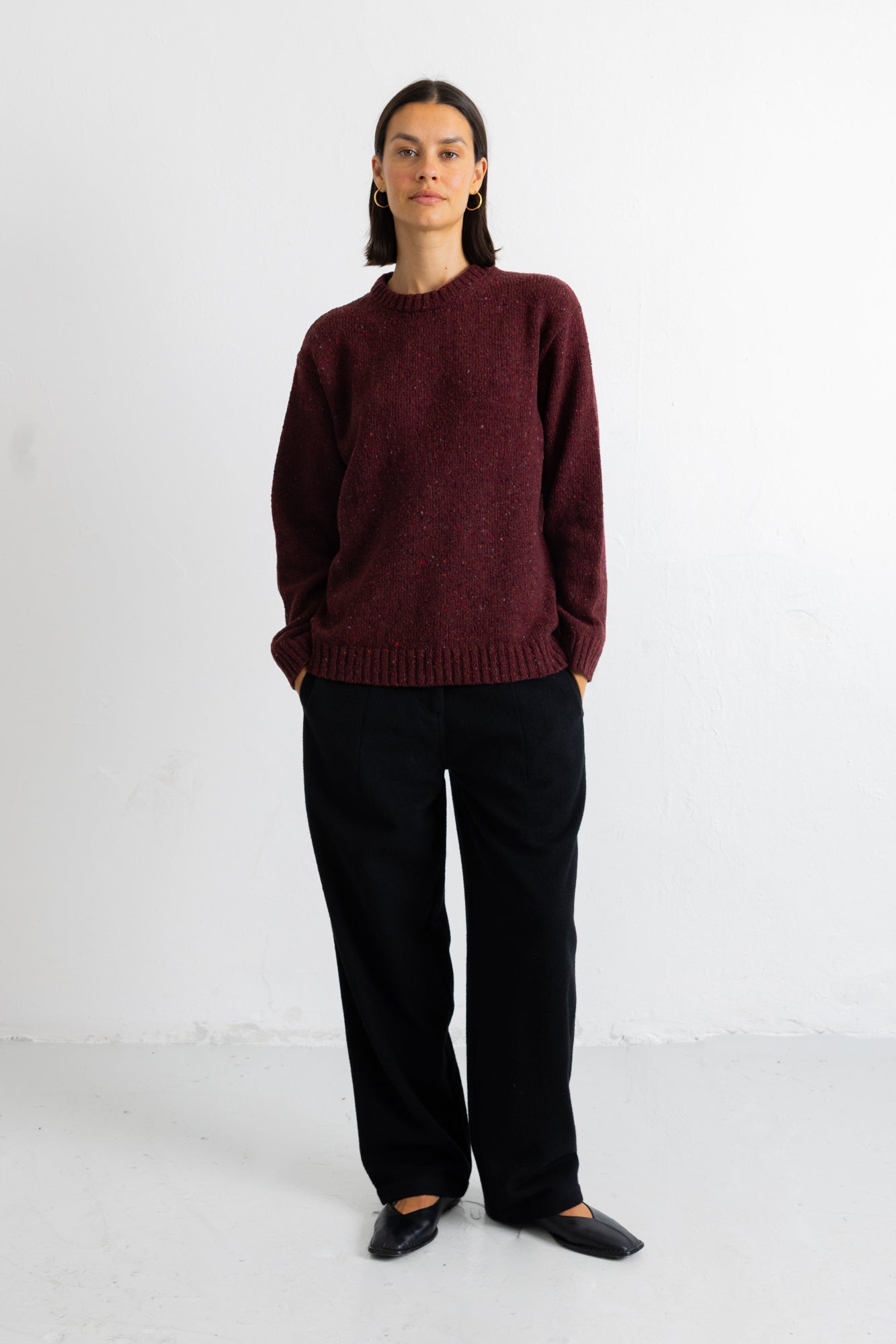 A woman stands against a plain white studio wall wearing a chunky knit crewneck sweater in deep burgundy, black wide-leg trousers, and black pointed flats.