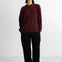 A woman stands against a plain white studio wall wearing a chunky knit crewneck sweater in deep burgundy, black wide-leg trousers, and black pointed flats.