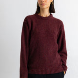 A woman wearing a textured, heathered burgundy knit crewneck sweater and dark trousers, standing against a plain white wall.