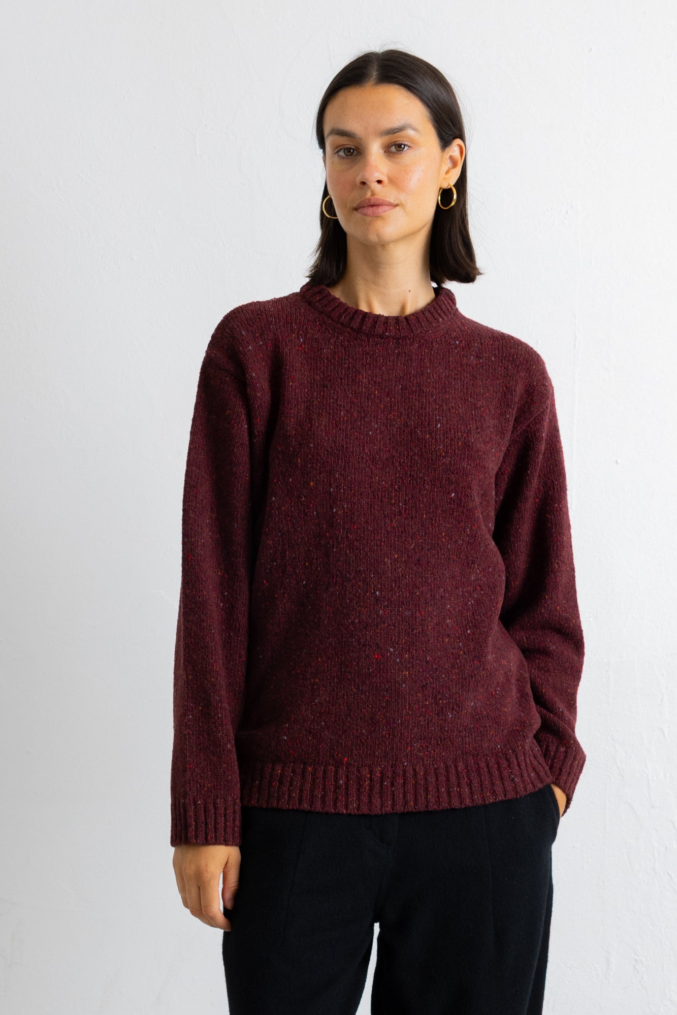 A woman wearing a textured, heathered burgundy knit crewneck sweater and dark trousers, standing against a plain white wall.