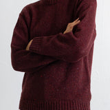 A person wearing a maroon knit crewneck sweater with a heathered texture, paired with black pants, against a plain white background.