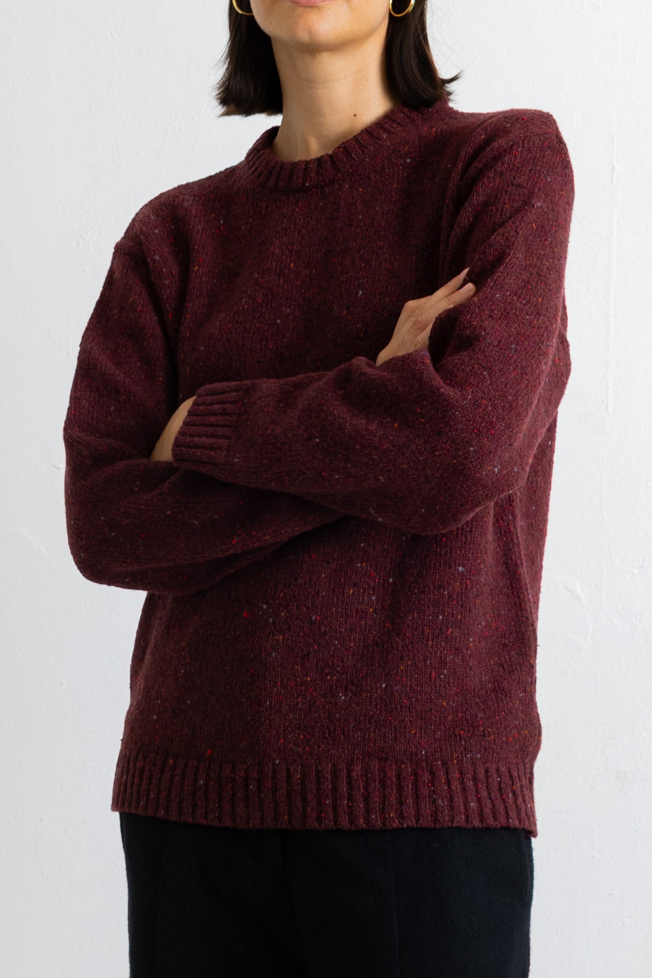 A person wearing a maroon knit crewneck sweater with a heathered texture, paired with black pants, against a plain white background.