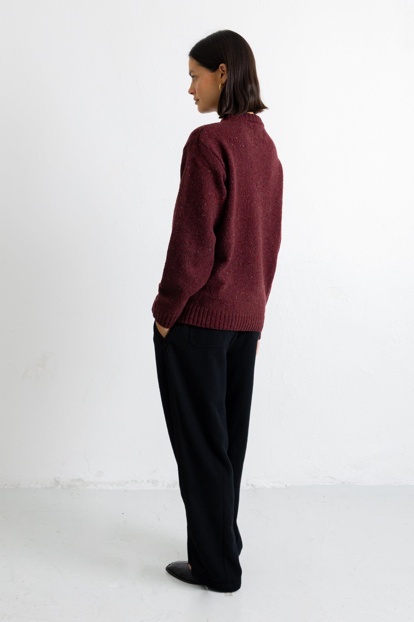 A model is shown from the back wearing a maroon knit sweater and black trousers, standing against a plain white studio background.