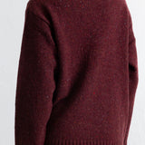 A person shown from the back wearing a dark burgundy knit sweater with a high ribbed collar, long sleeves, and a ribbed hem, paired with black pants against a white wall.