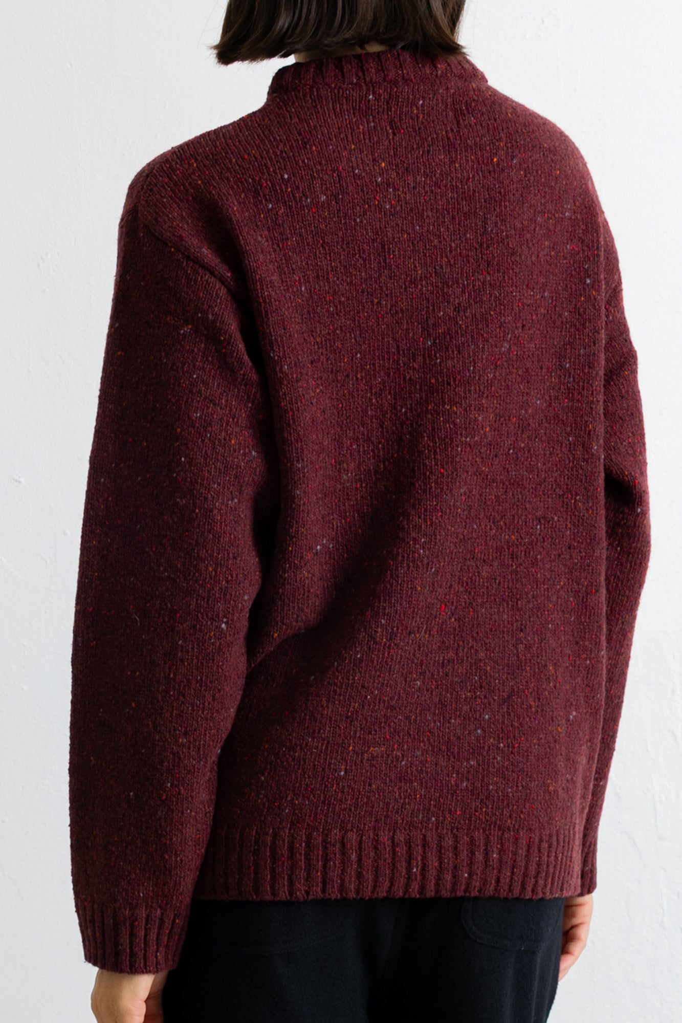 A person shown from the back wearing a dark burgundy knit sweater with a high ribbed collar, long sleeves, and a ribbed hem, paired with black pants against a white wall.