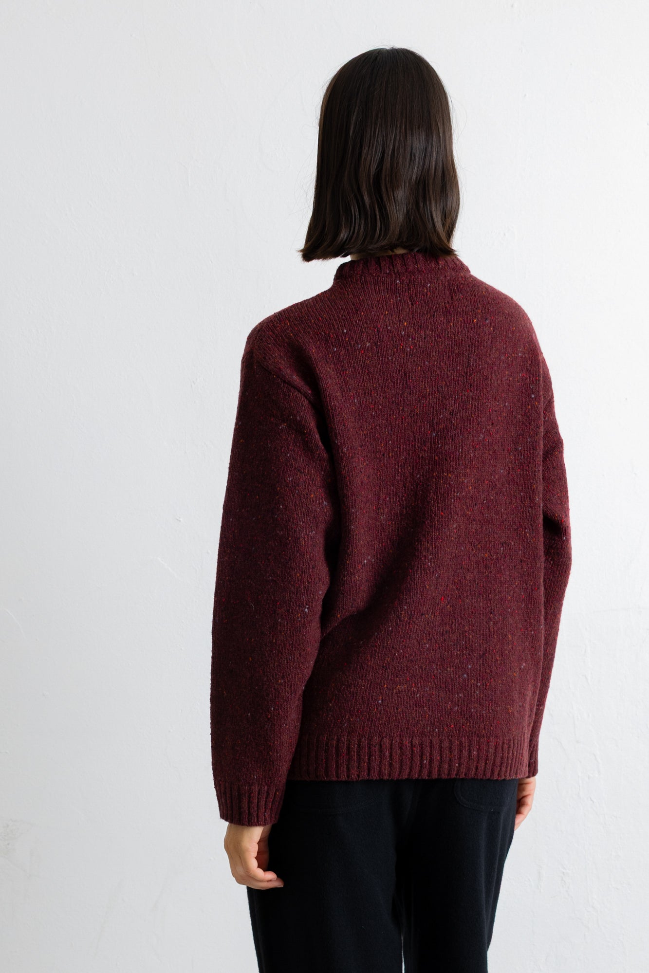 A person shown from the back wearing a dark burgundy knit sweater and black pants against a plain white wall.