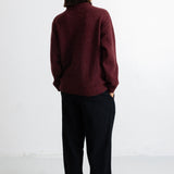A person shown from the back wearing a dark burgundy knit sweater and black wide-leg trousers, standing against a plain white studio background.