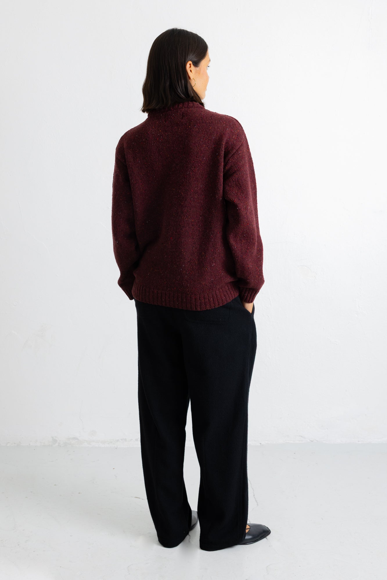 A person shown from the back wearing a dark burgundy knit sweater and black wide-leg trousers, standing against a plain white studio background.