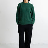 A woman stands against a plain white studio wall wearing a dark green knit sweater, black wide-leg trousers, and black flats.