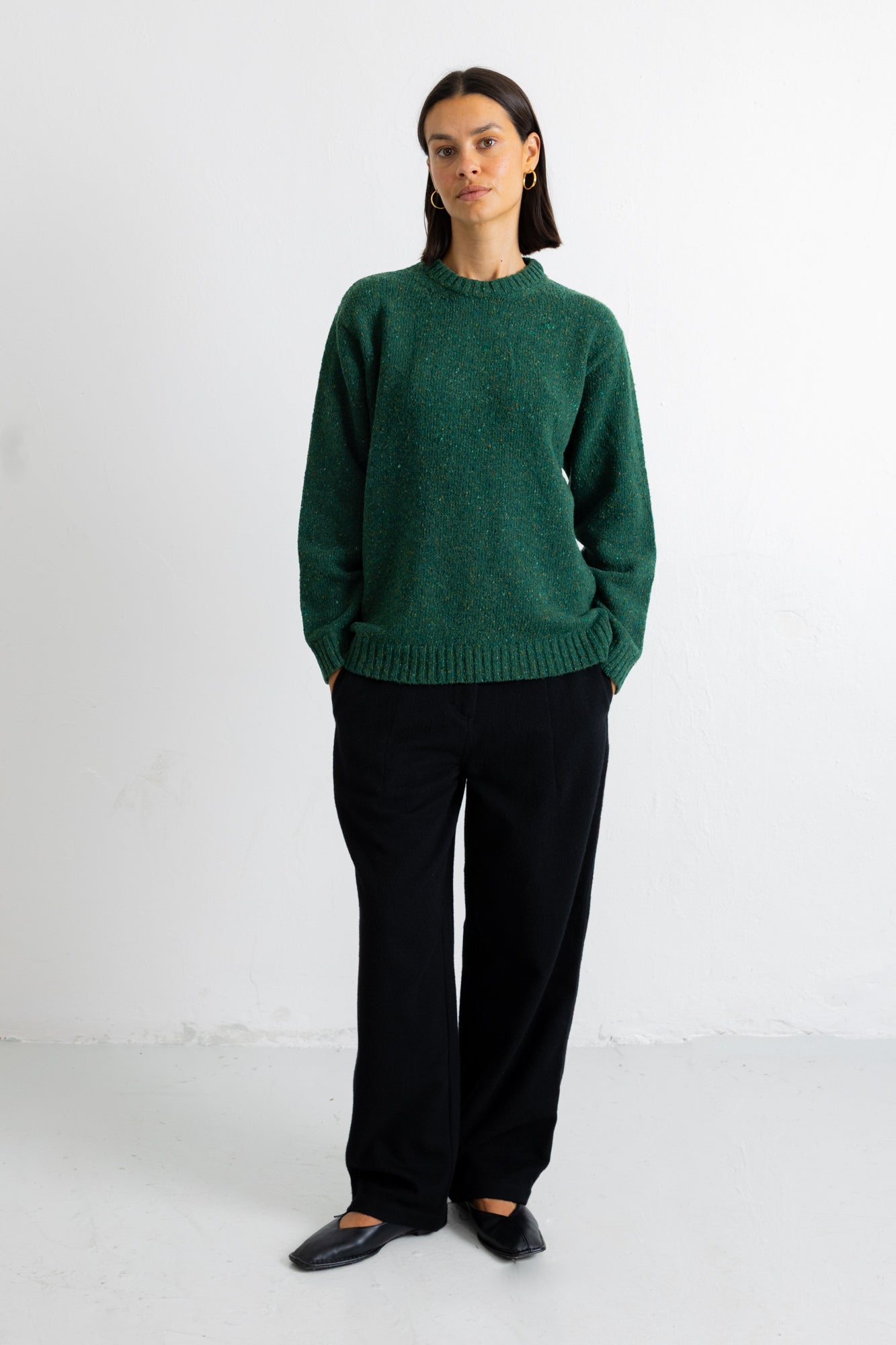 A woman stands against a plain white studio wall wearing a dark green knit sweater, black wide-leg trousers, and black flats.