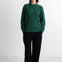 A woman stands against a plain white studio wall wearing a dark green knit sweater, black wide-leg trousers, and black flats.